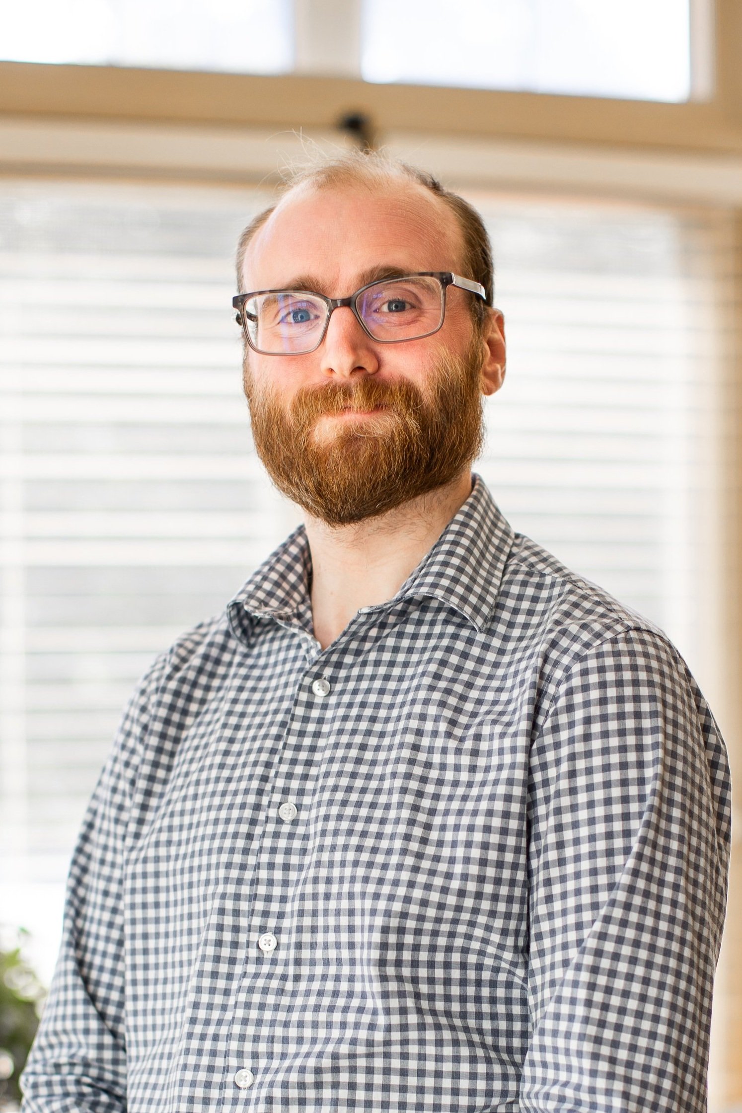 A man with glasses and a beard wearing a checkered button-up shirt, standing indoors with a window in the background.