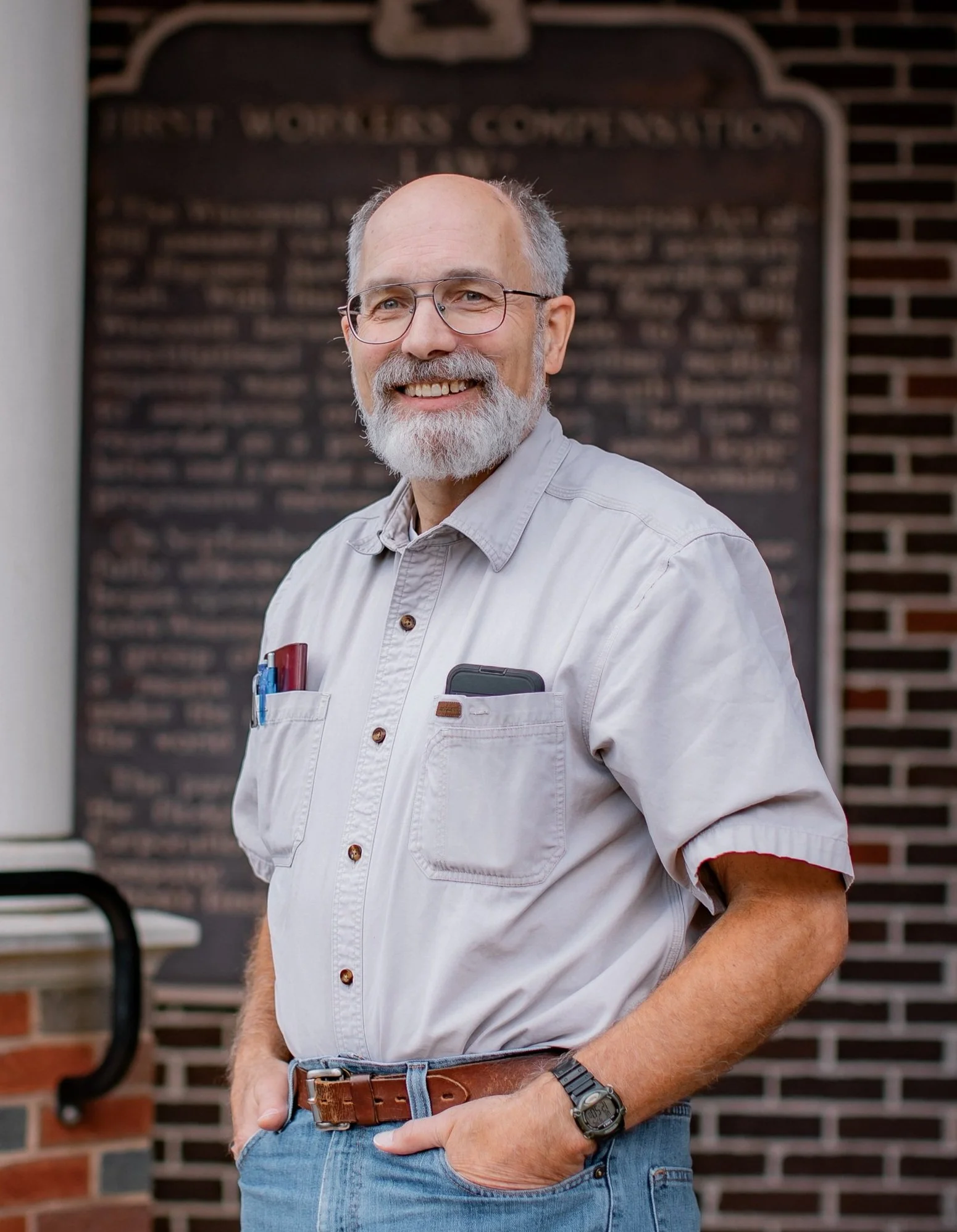 A smiling older man with glasses and a gray beard standing outdoors in front of a chalkboard with writing. He is wearing a light-colored button-up shirt, blue jeans, and a wristwatch, with pens in his shirt pocket and his hand in his pocket.