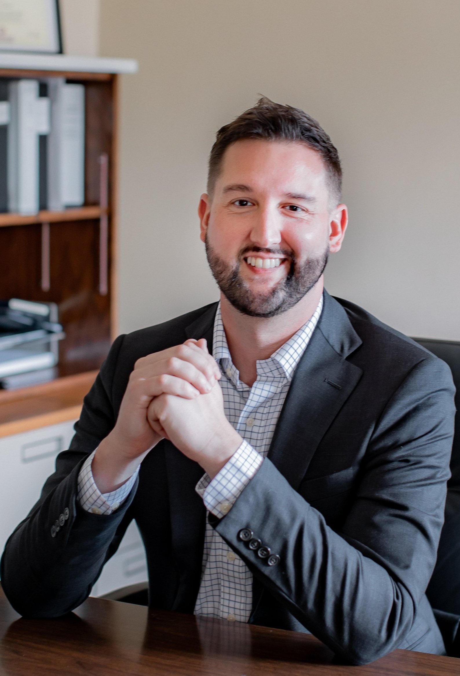 A smiling man in a suit sitting at a desk with hands clasped, in an office setting.