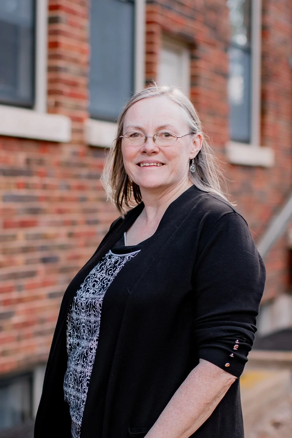 A woman with glasses and earrings, wearing a black jacket and a patterned blouse, standing outdoors in front of a brick building with windows.