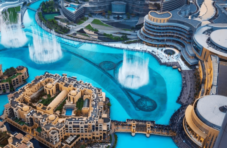 Aerial view of a large fountain with water jets surrounded by modern architecture and buildings with pools and terraces.