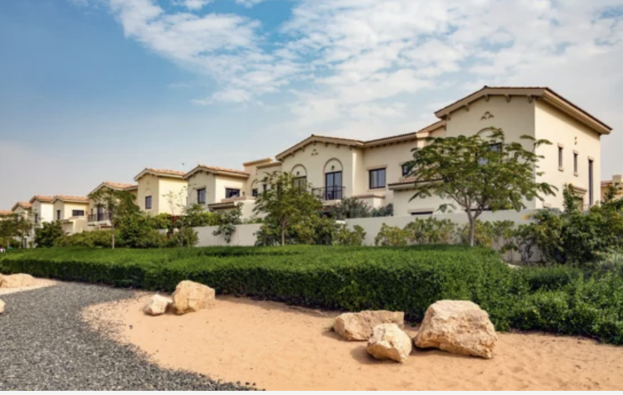Row of Mediterranean-style villas with landscaped gardens surrounded by shrubs and trees under a blue sky.