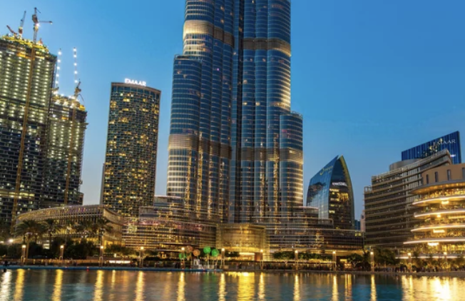 Skyline with illuminated skyscrapers at dusk, including a tall, iconic building, reflecting on a waterfront.