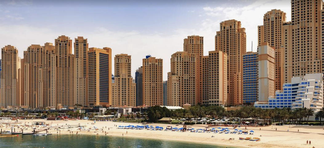 Skyline of high-rise buildings along a sandy beach with umbrellas and sunbathers, under a partly cloudy sky."