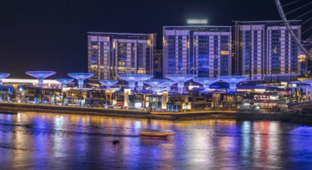 Night view of a waterfront cityscape with illuminated buildings and abstract structures, reflecting on the water.