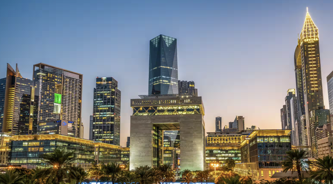 Modern skyline of a city with illuminated skyscrapers, including a distinctive arch-shaped building and tall towers at dusk.