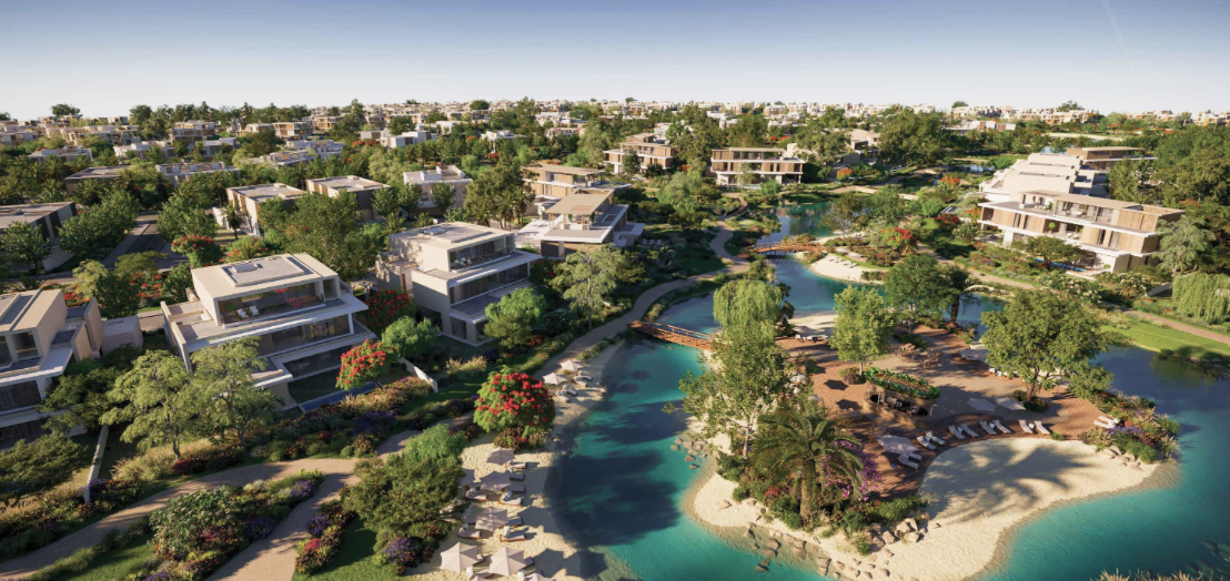 Aerial view of a luxurious residential community featuring modern villas, lush greenery, and landscaped gardens surrounding a central waterway with a sandy beach area and wooden pathways.