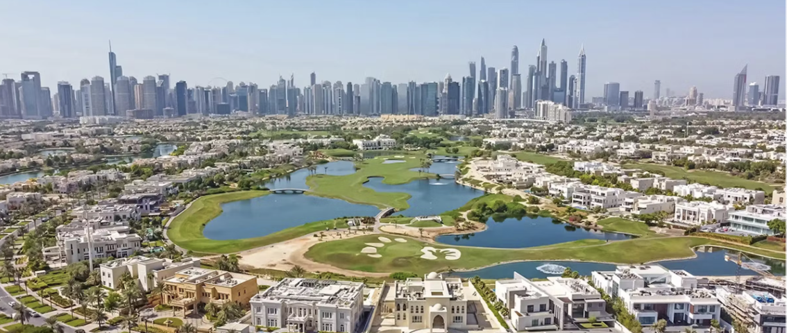 Aerial view of a golf course surrounded by luxury villas with a city skyline in the background.
