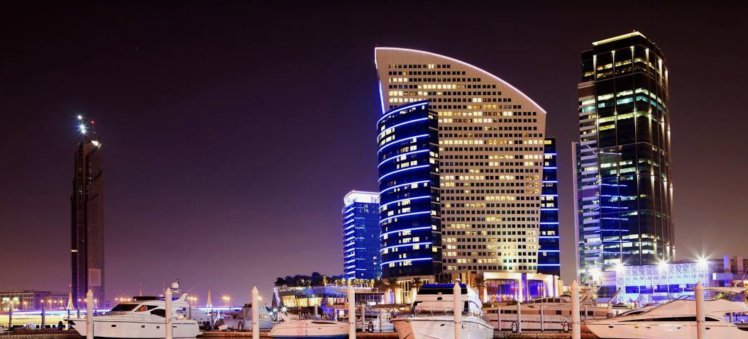 Night view of modern city skyline with illuminated skyscrapers and moored yachts in the foreground.