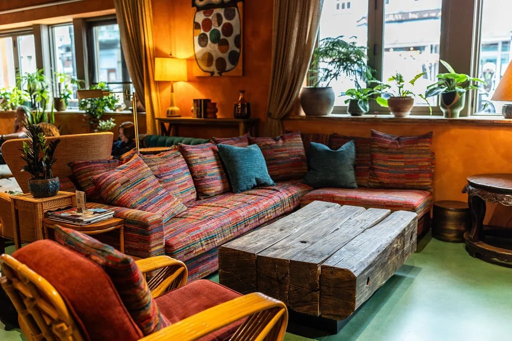 A cozy living room with a colorful striped couch and matching armchair, surrounded by potted plants on the windowsill, a rustic wooden coffee table, and a side table with magazines and a plant. The room has warm lighting and large windows with curtai