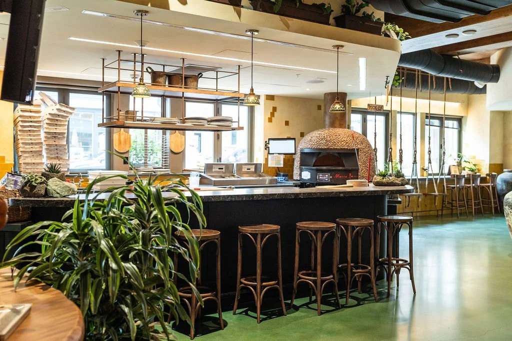 Interior of a restaurant with a pizza oven, bar seating, and large windows.