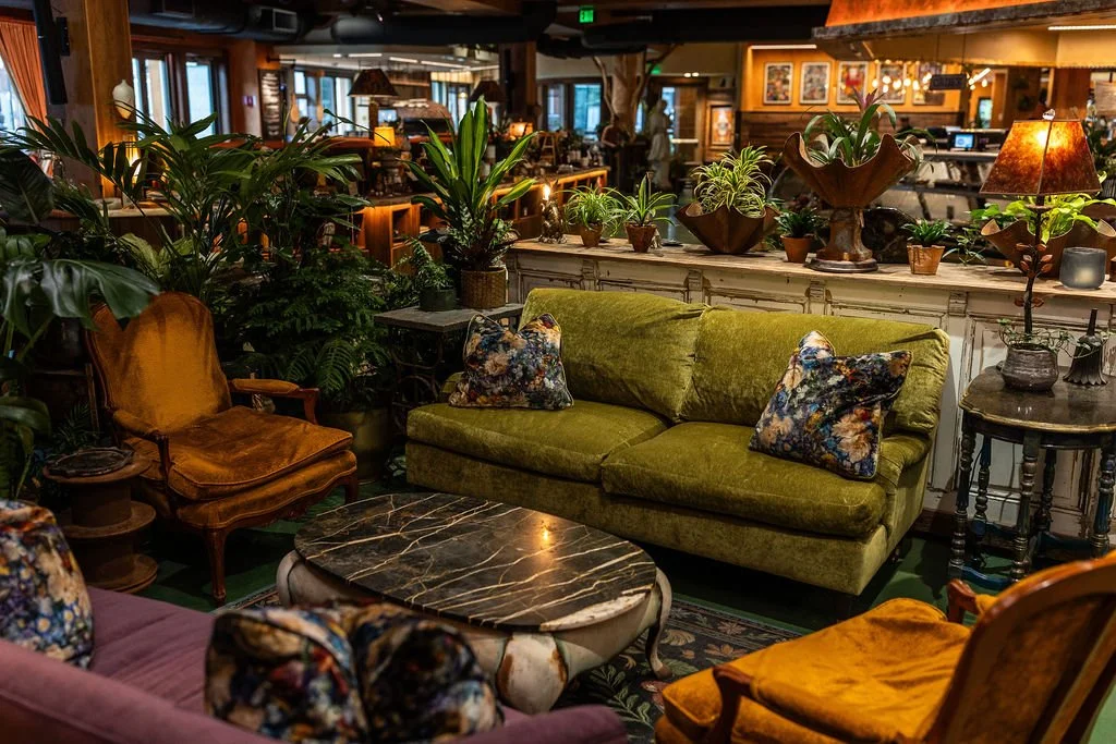 A cozy indoor lounge area with green velvet sofa, vintage yellow armchairs, floral cushions, a black marble coffee table, and numerous potted plants, with warm lighting and wooden decor.