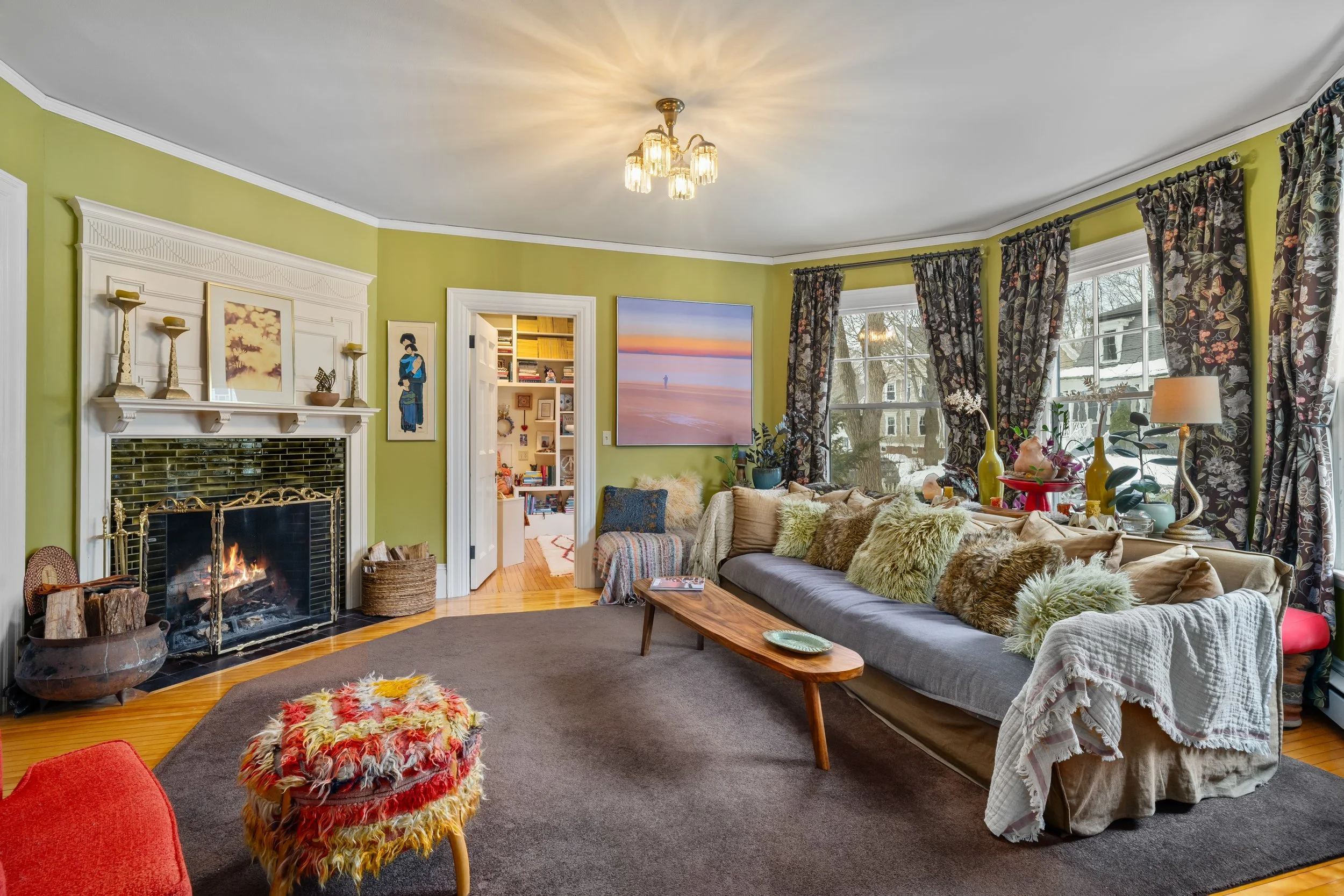 Living room with green walls, fireplace with books and decorations, beige sofa with fuzzy pillows, a wooden table, large windows with floral curtains, and various decorative items on the windowsill.