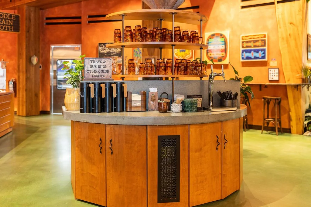 A cafe or restaurant counter with stacked orange glasses on a rotating shelf, black water bottles, a sink, and various containers. The background features wooden walls, colorful art, and seating areas.
