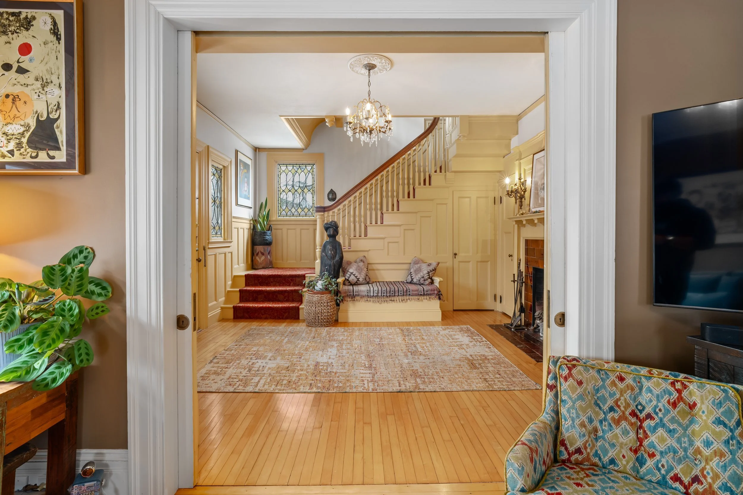 Living room with a view of a staircase, fireplace, and seating area, decorated in vintage style with a chandelier, artwork, plants, and a patterned rug.