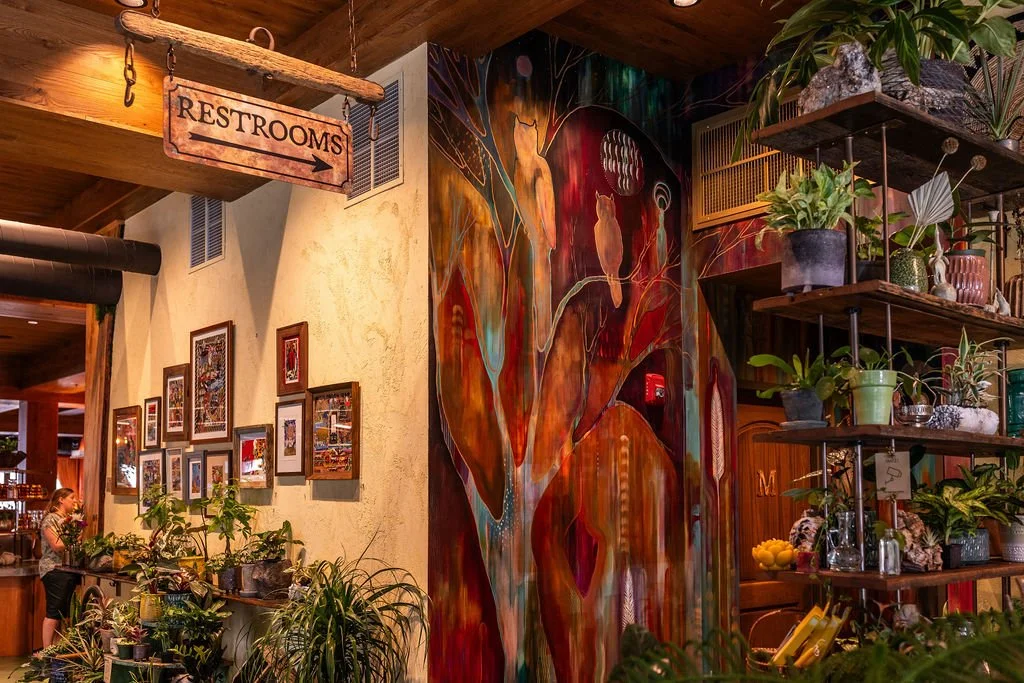 Interior of a restaurant or cafe with a colorful abstract mural of a tree and animals, potted plants on shelves, framed pictures on the wall, and a wooden ceiling with a sign pointing to restrooms.