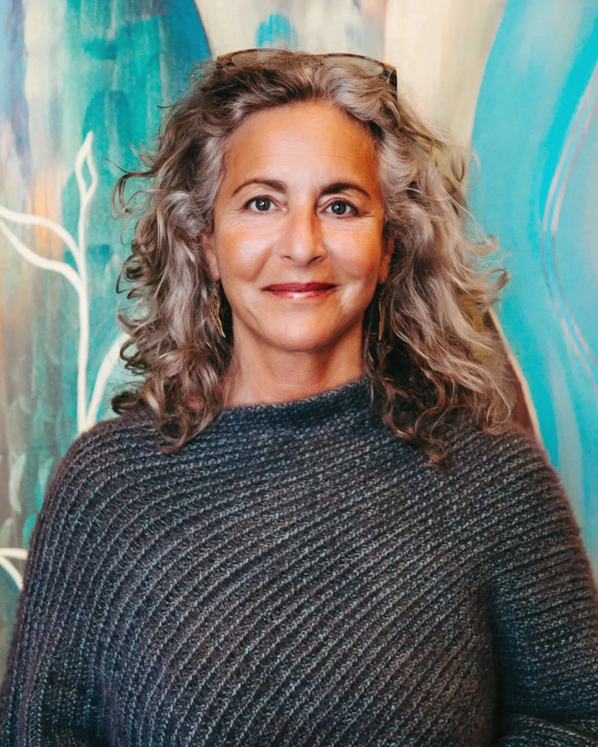 A woman with shoulder-length curly gray hair, wearing a dark textured sweater, standing in front of a colorful abstract background.