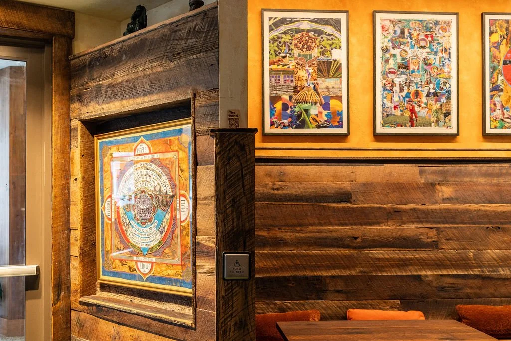 Decorative artwork on a wooden wall inside a restaurant or cafe with framed colorful paintings and a wooden table with orange seats.