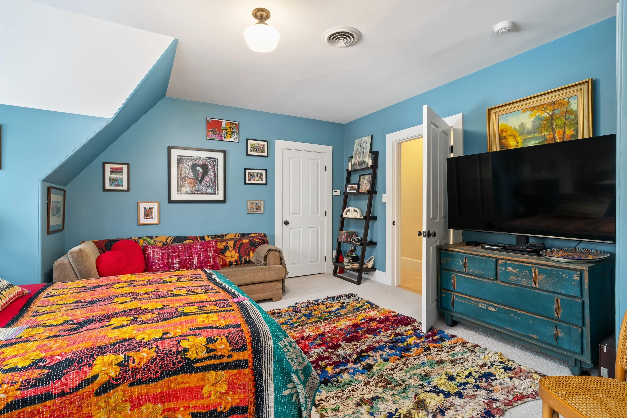 A colorful bedroom with blue walls, a bed with bright multicolored quilt, a beige sofa with red pillows, a blue dresser with a large flat-screen TV, picture frames on the wall, a ladder-style shelf, and a multicolored rug.