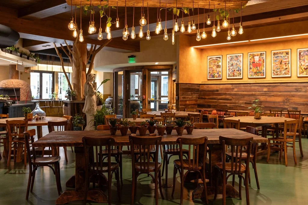 Interior of a cozy restaurant with wooden tables and chairs, art frames on a wall, hanging Edison bulbs, houseplants, and a tree with a statue nearby.