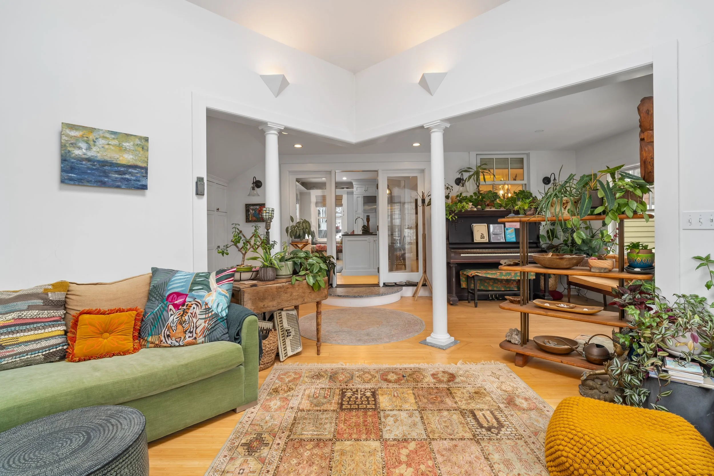 Living room with a green sofa, wooden shelves with plants, a black piano, an ornate area rug, and white walls with columns.