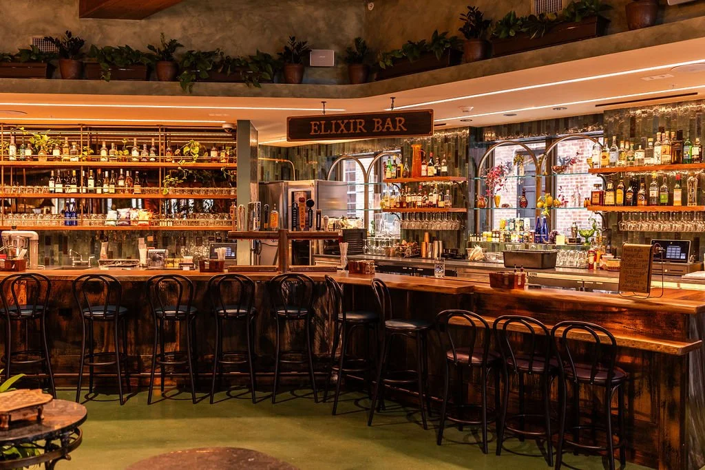 Bar with a wooden counter, black chairs, and shelves stocked with liquor bottles, green plants, and a sign that reads 'Elixir Bar' in a cozy, warmly lit setting.