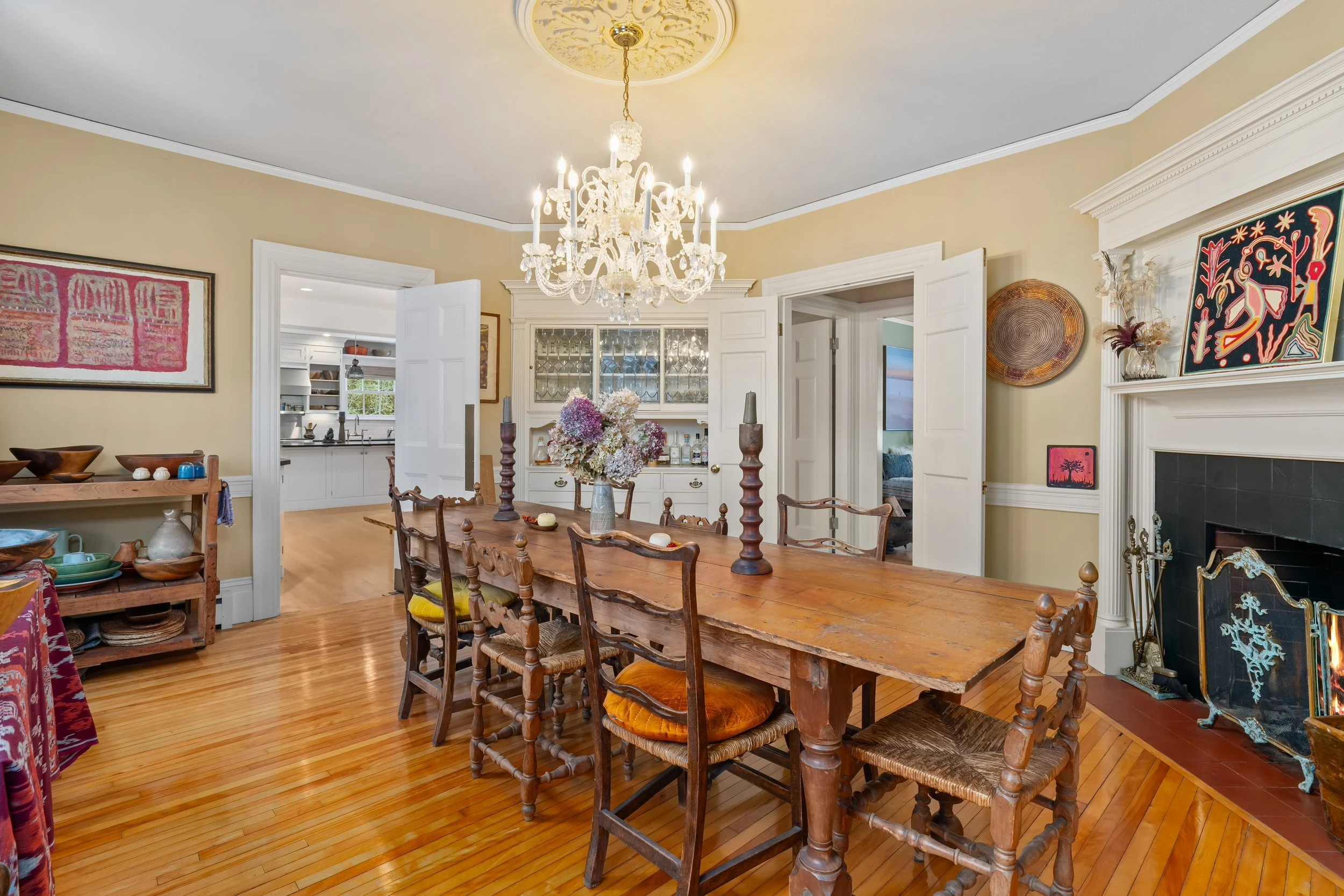 A dining room with a rustic wooden table and six matching chairs, a chandelier hanging from the ceiling, a fireplace with art above, and a buffet with dishes, with a view into the kitchen and an additional room.