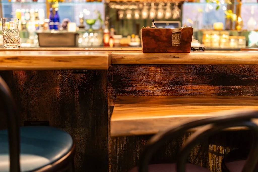 Interior of a bar or restaurant with a polished wooden counter, a small wooden napkin holder, and seating with dark chairs in the foreground.