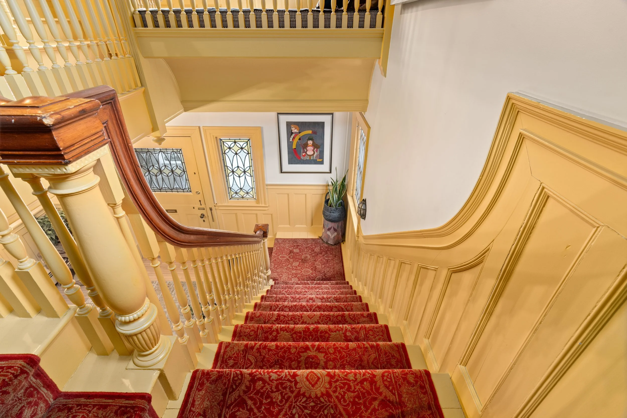 View of a staircase with red carpet, leading to a front door with decorative glass panels. Yellow wall panels and a framed abstract artwork hang on the wall, with a potted plant near the door.