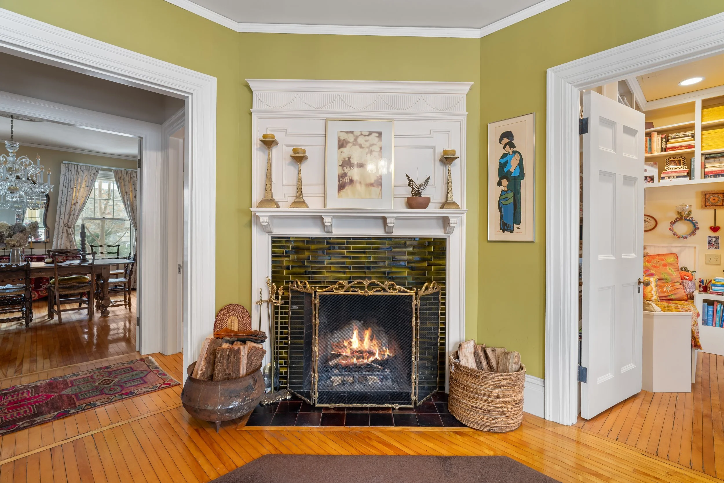 Living room with a lit fireplace, green walls, and decorative objects on the white mantle. On the left, there is a doorway leading to a dining room, and on the right, another doorway leading to a room with built-in shelves and a chair. Wood flooring