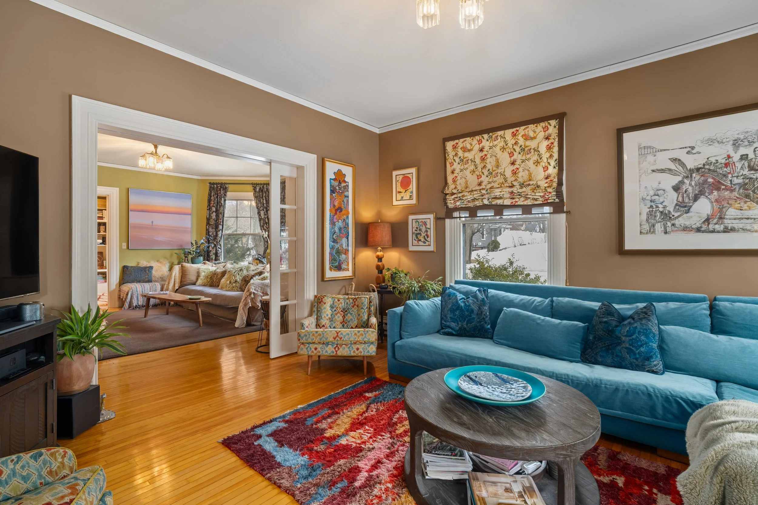 Living room with blue sofa, colorful artwork, and window with patterned valance. Adjacent room with sofa, paintings, and curtains visible through open doorway.