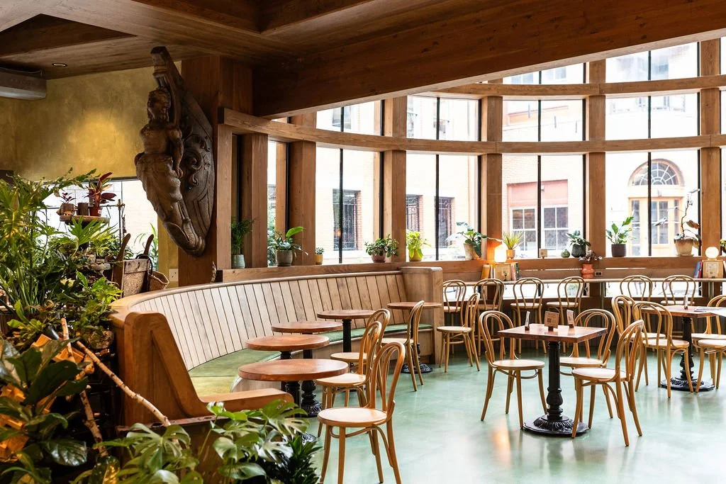 Interior of a cozy cafe with wooden furniture, large curved windows, potted plants, and green flooring.