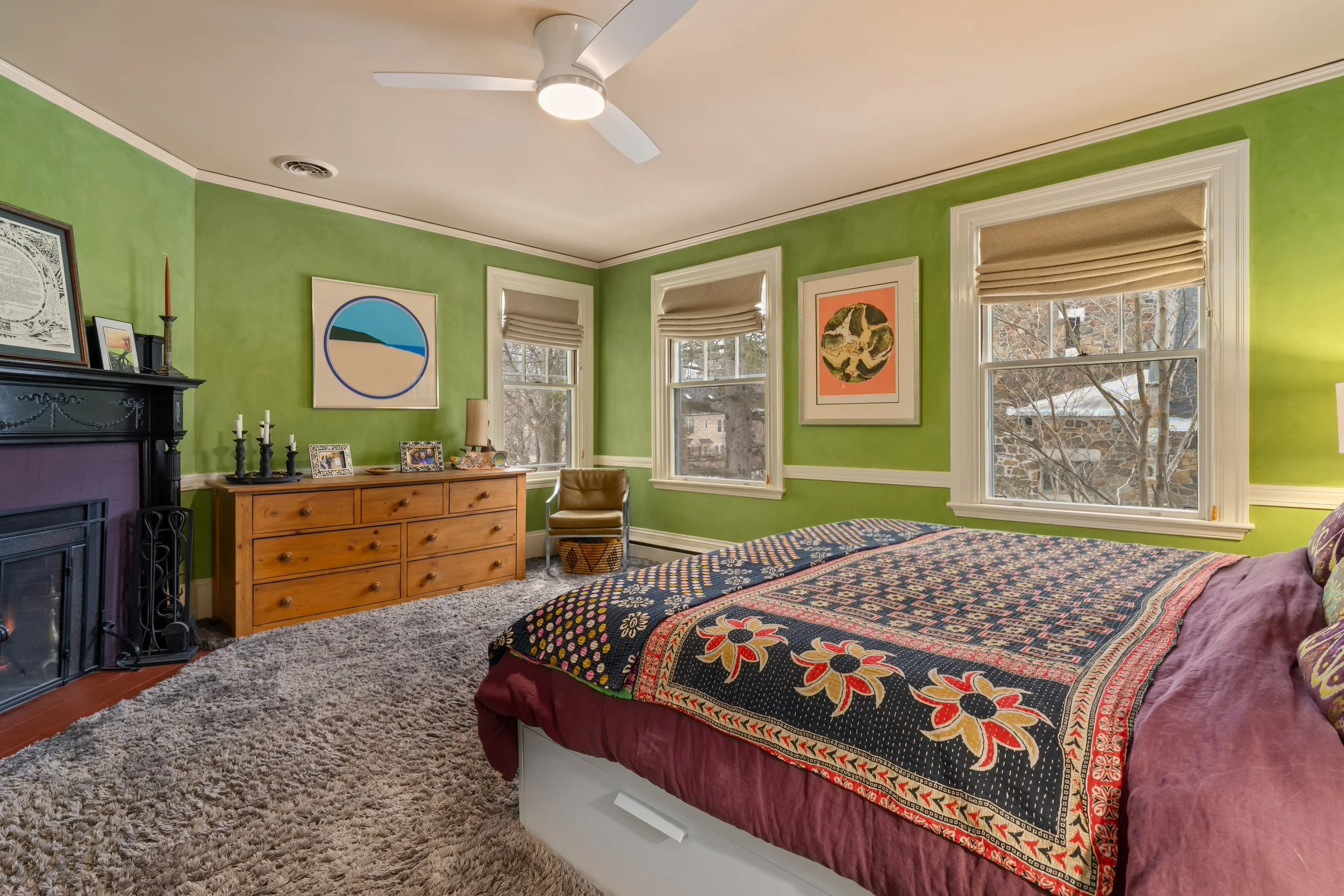 A bedroom painted in bright green with three windows showing trees outside. The room has a black fireplace, a wooden dresser with framed photos and candles, and a bed with a colorful patterned quilt and maroon bedding. A tan chair and a textured beig