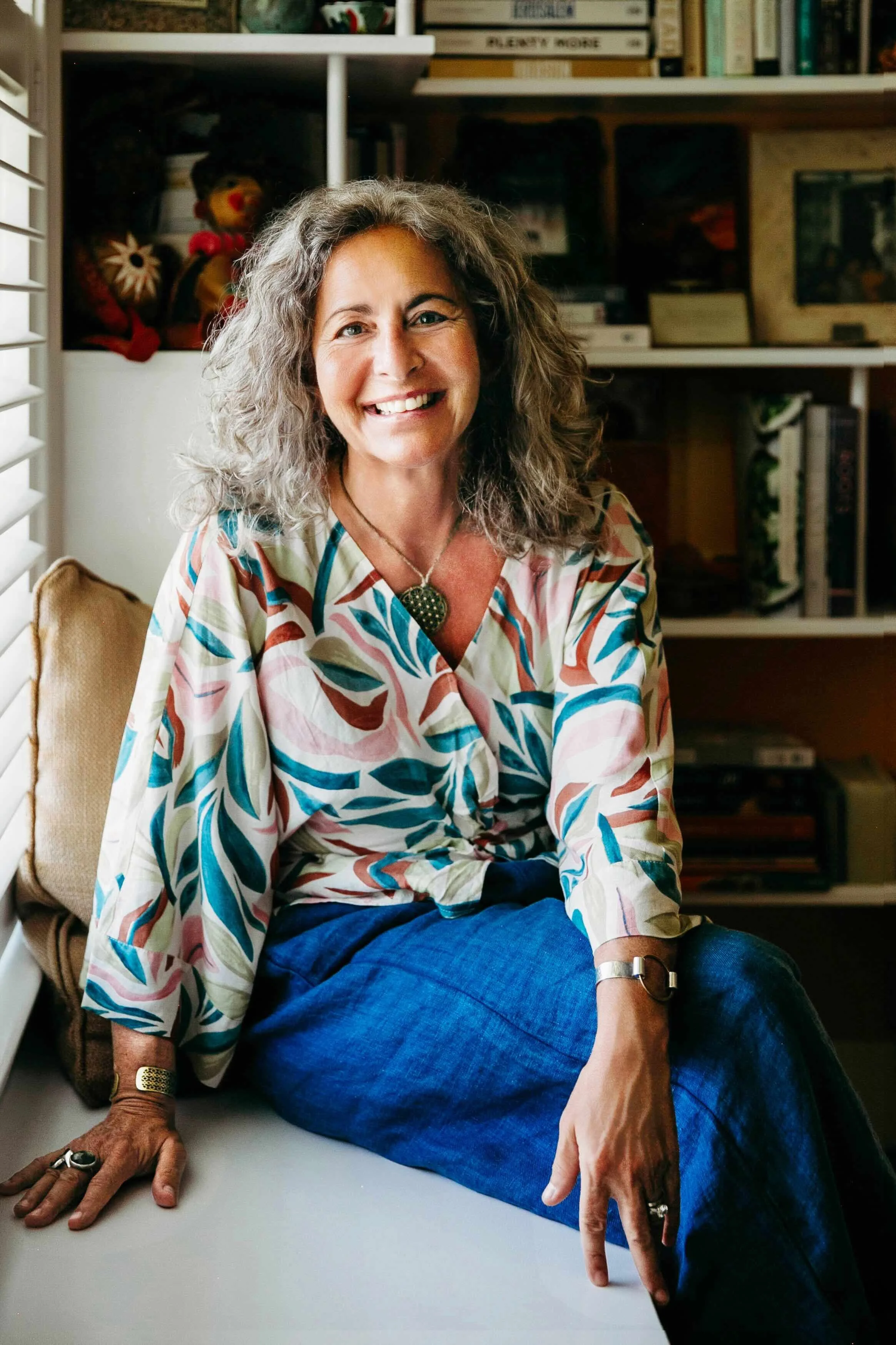 A woman with curly gray hair, smiling at the camera, sitting on a white surface in a room with a bookshelf and window blinds visible in the background. She is wearing a colorful patterned blouse, blue pants, and jewelry, including rings, a bracelet, and a necklace.