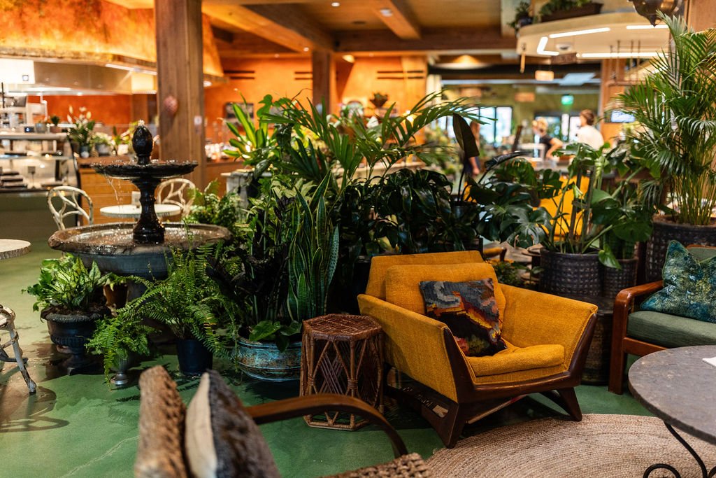 Interior of a cozy cafe or restaurant with lush green plants, a mustard yellow armchair with a decorative pillow, and wooden furniture. There is a multi-tiered water fountain on the left side and people working or dining in the background.