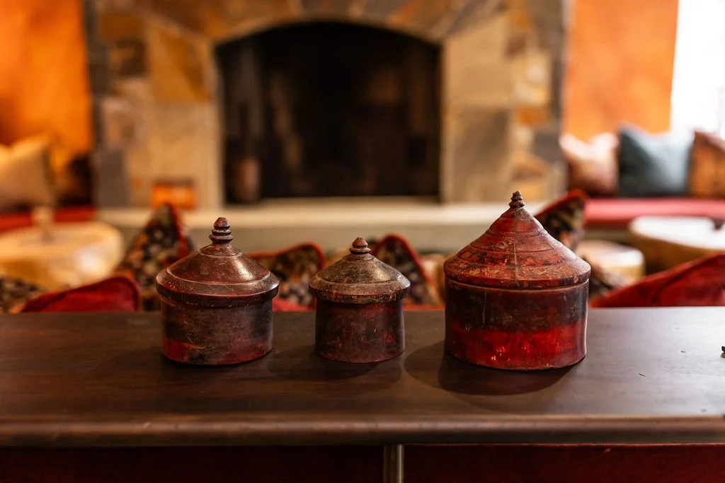 Three antique metal containers with pointed lids on a wooden table in front of a cozy fireplace with a stone and wood surround.