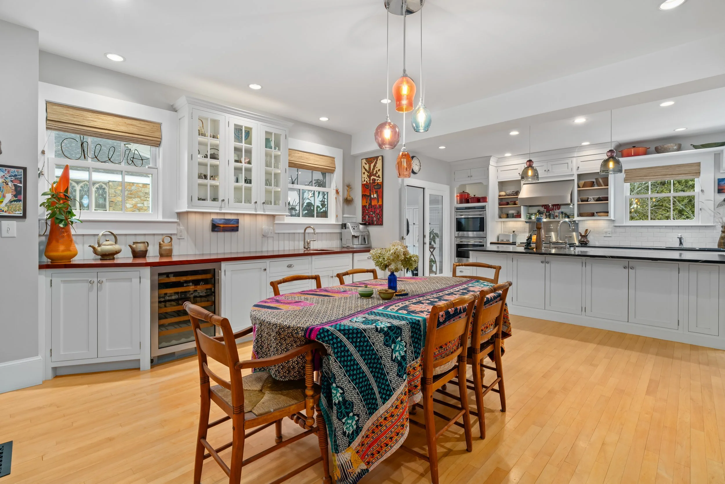 Bright kitchen with white cabinets, wooden flooring, and colorful pendant lights over a dining table with a patterned tablecloth. The table has wooden chairs, a blue vase with flowers, and small bowls. The kitchen has windows with blinds and various