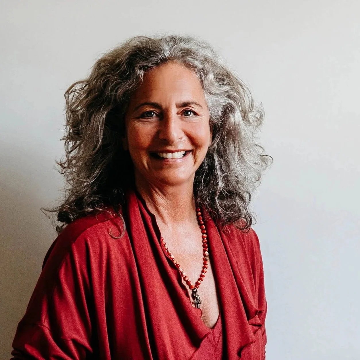 A smiling woman with curly gray hair wearing a red blouse and necklace, standing against a plain light-colored background.