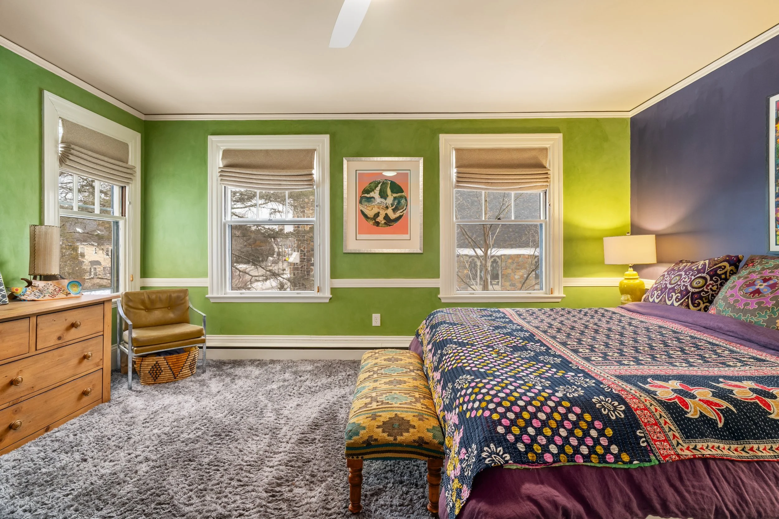 A bedroom with green and purple walls, three windows with beige blinds, a bed with a colorful quilt, a yellow lamp, a wooden dresser, a tan chair, and a patterned pouf at the foot of the bed.