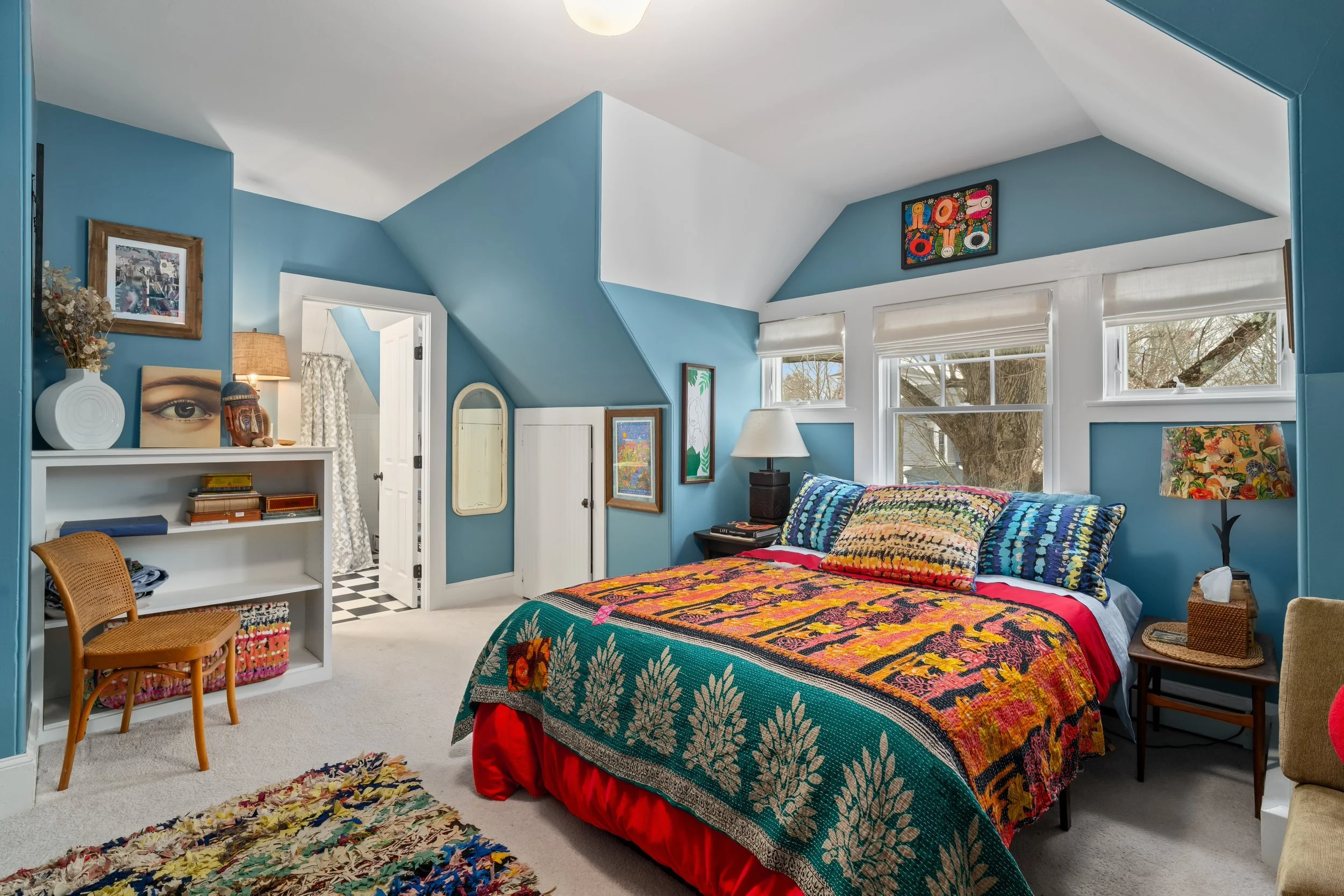 Colorful bedroom with blue walls, a bed with vibrant quilt and pillows, and a window showing trees outside.