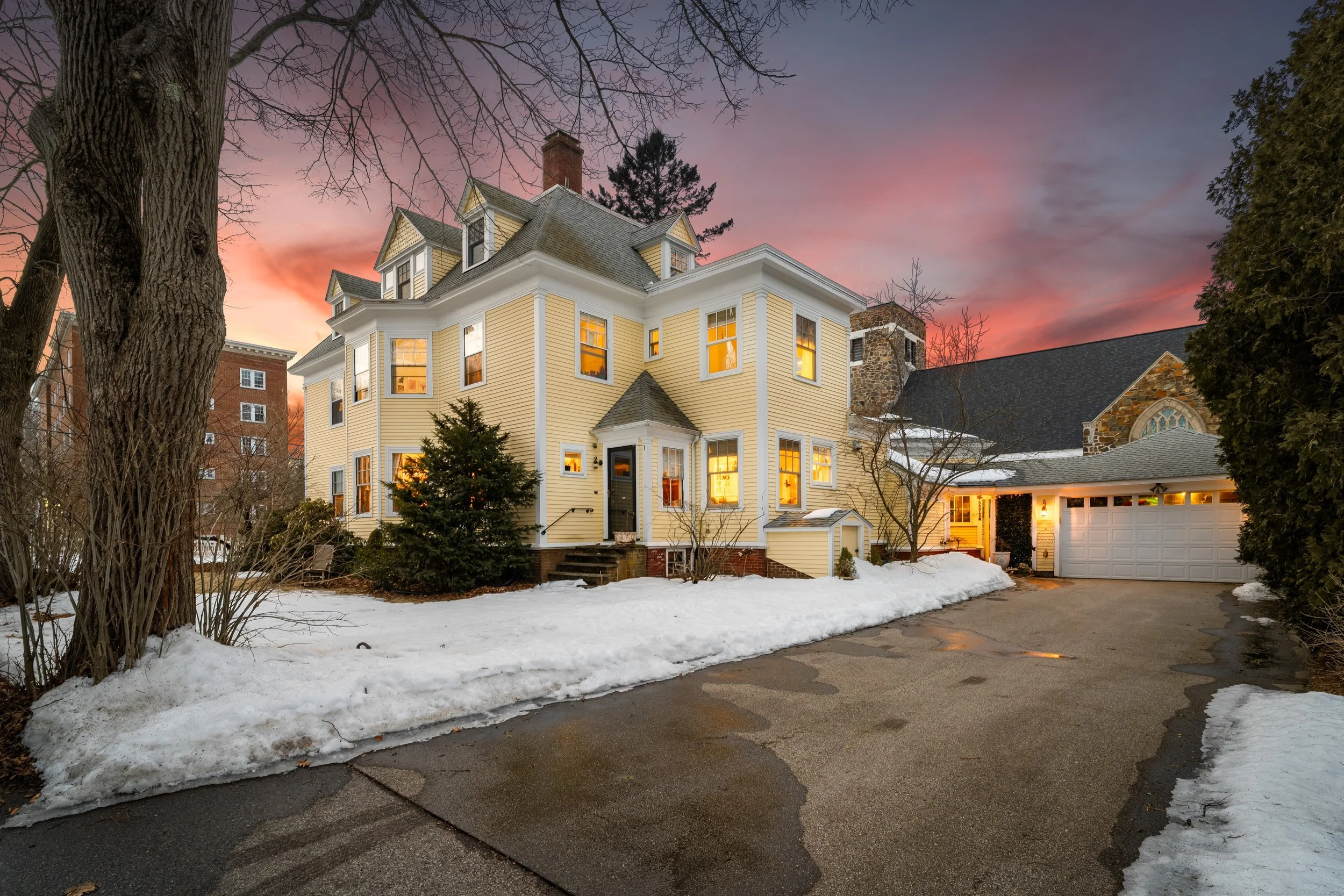 A large, historic house with yellow siding and multiple gables, illuminated from inside, during sunset in winter. Snow covers the ground, the driveway is wet with patches of snow, and leafless trees frame the scene.
