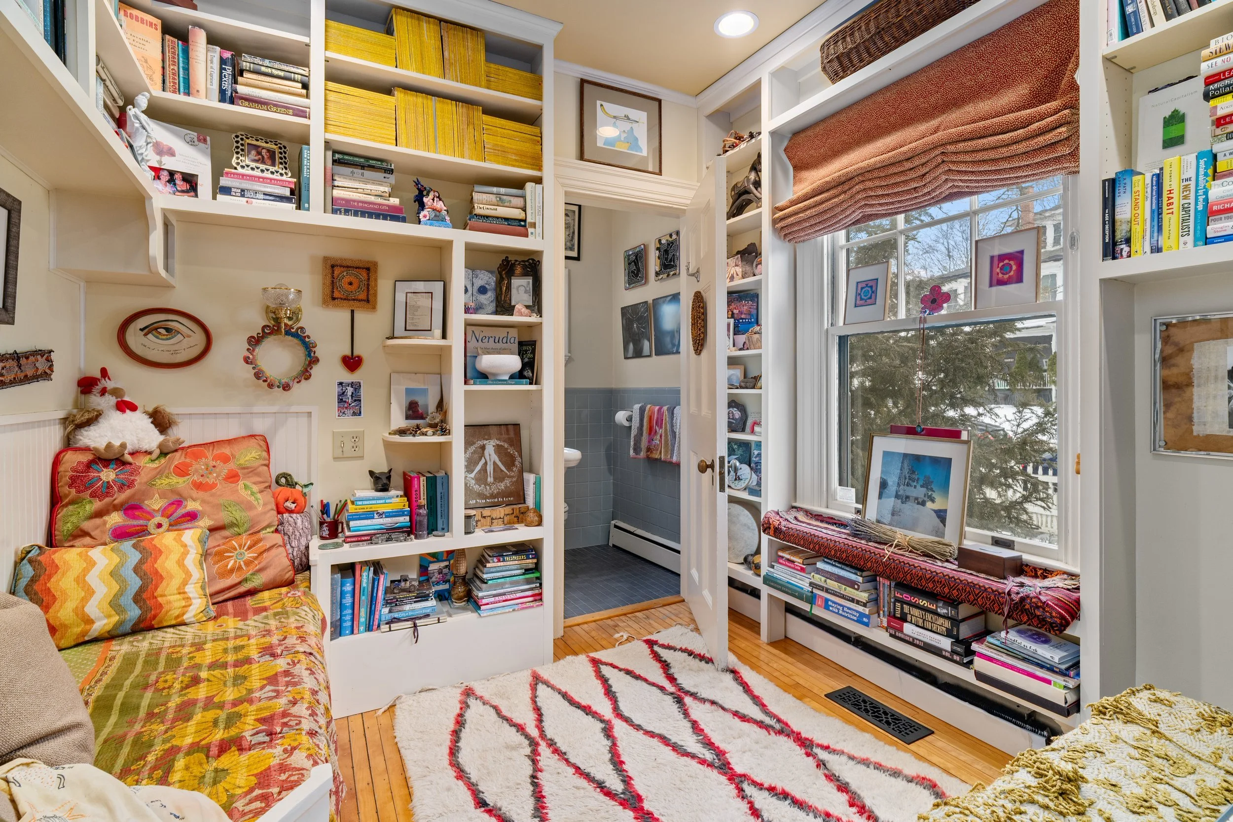 A cozy living room with white built-in bookshelves filled with books and decorative items. A window with pink curtains allows natural light. A colorful rug covers the hardwood floor. An open door reveals a small bathroom with blue tiles. The room is