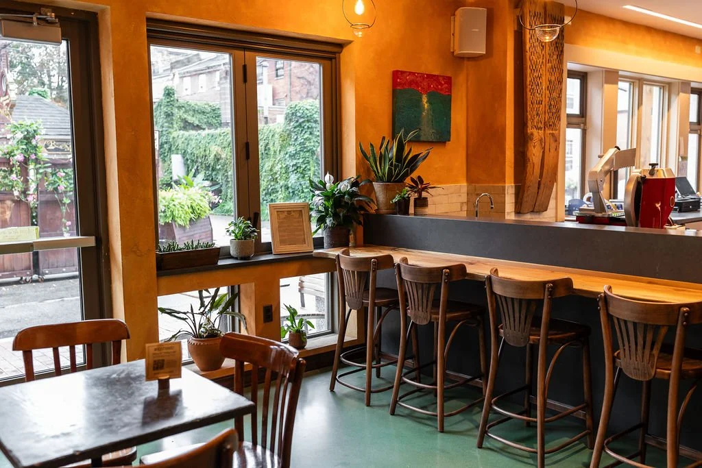 Interior of a cozy cafe with wooden chairs, tables, and a bar area with high chairs. Large windows reveal a green outdoor space with plants and trees.