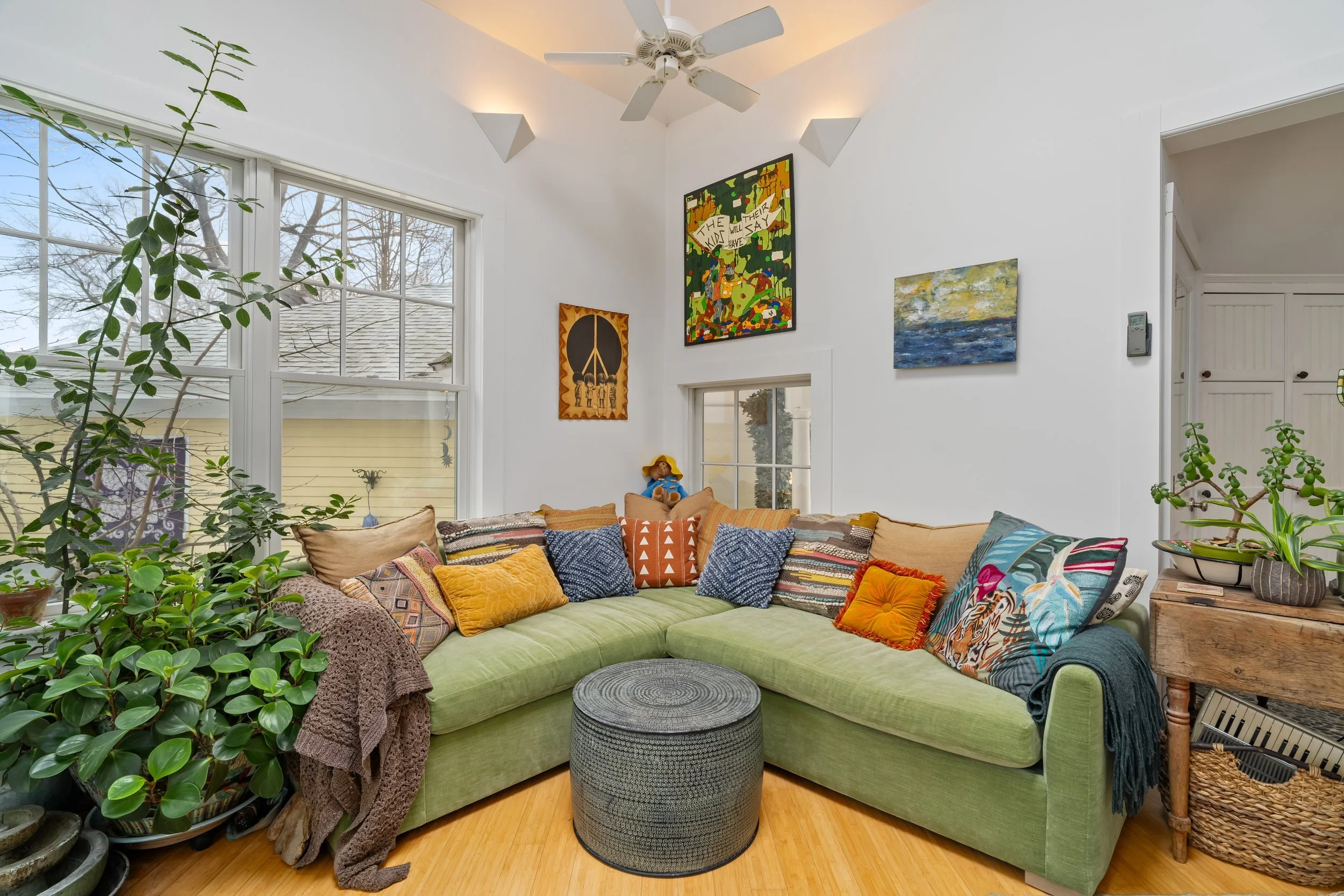 A living room with a green sectional sofa filled with colorful pillows, large windows, and wall art including paintings and a hanging decorative piece. There are plants, a small black ottoman in front of the sofa, and a rustic wooden side table with