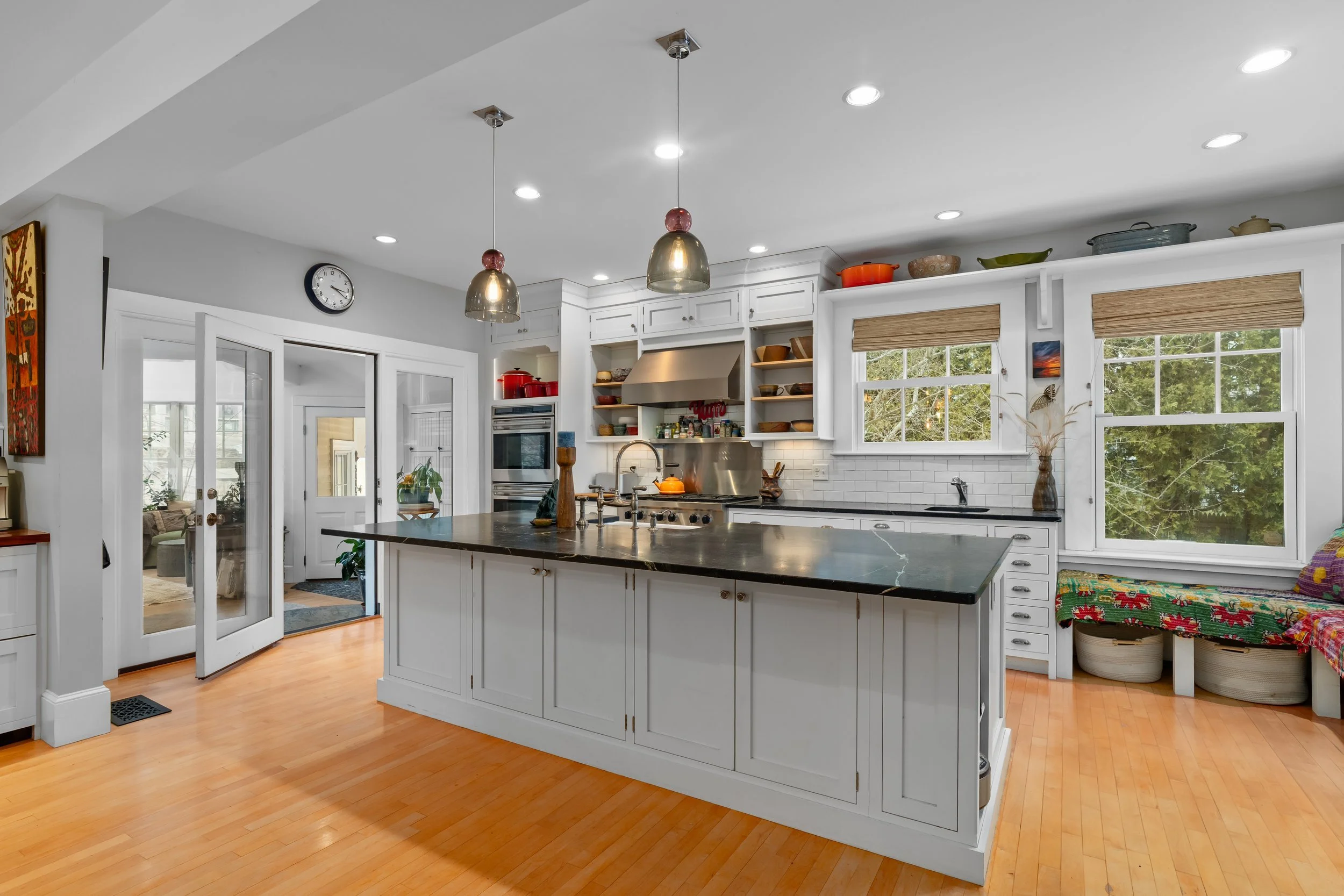 Bright kitchen with white cabinets, black countertops, and hardwood floors; open shelving with colorful dishes; large window overlooking greenery; kitchen island with sink and faucet; appliances including stainless steel oven and range hood; pendant