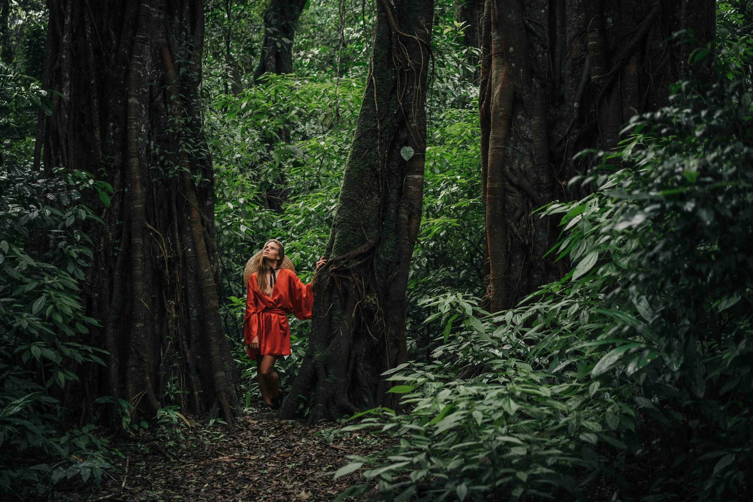 A woman in a red dress walking through a dense, green jungle with tall trees and thick foliage.