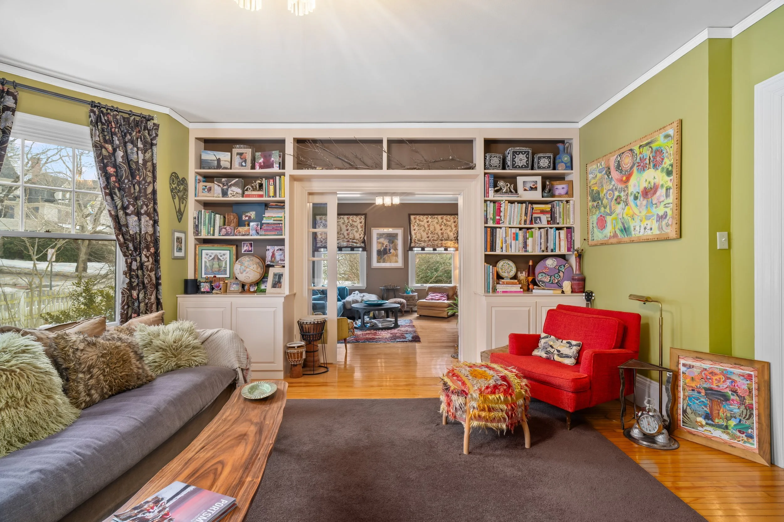 Living room with green walls, large window, beige built-in bookshelves filled with books, pictures, and decorative items, red armchair, beige sofa with green and brown pillows, wooden coffee table, artwork on walls, and a view into a second sitting a