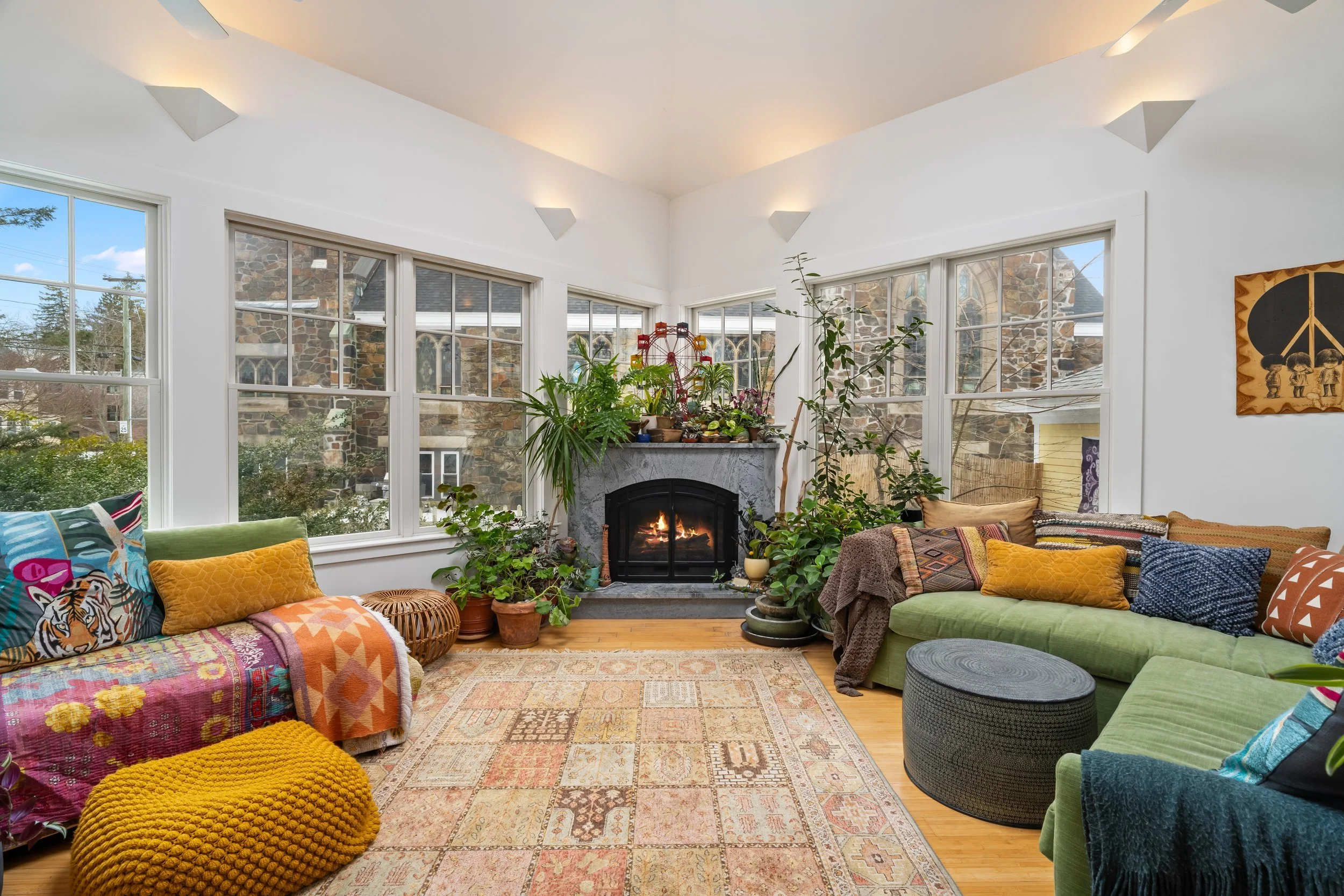 Bright living room with large windows, a fireplace with a fire, green and patterned sofas, colorful cushions and knitted poufs, plants near the fireplace, and a decorative wall hanging.