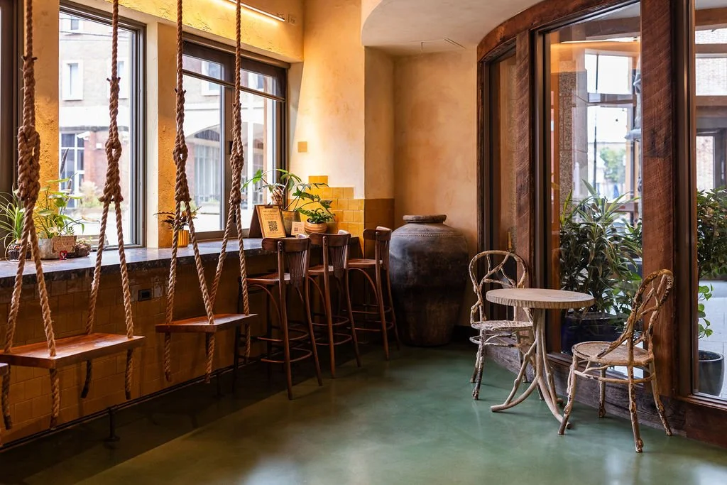 Interior of a cozy café or lounge with large windows, wooden furniture, a window seat with plants, and a small round table with two chairs near a glass door. Rope accents are hanging from the ceiling.