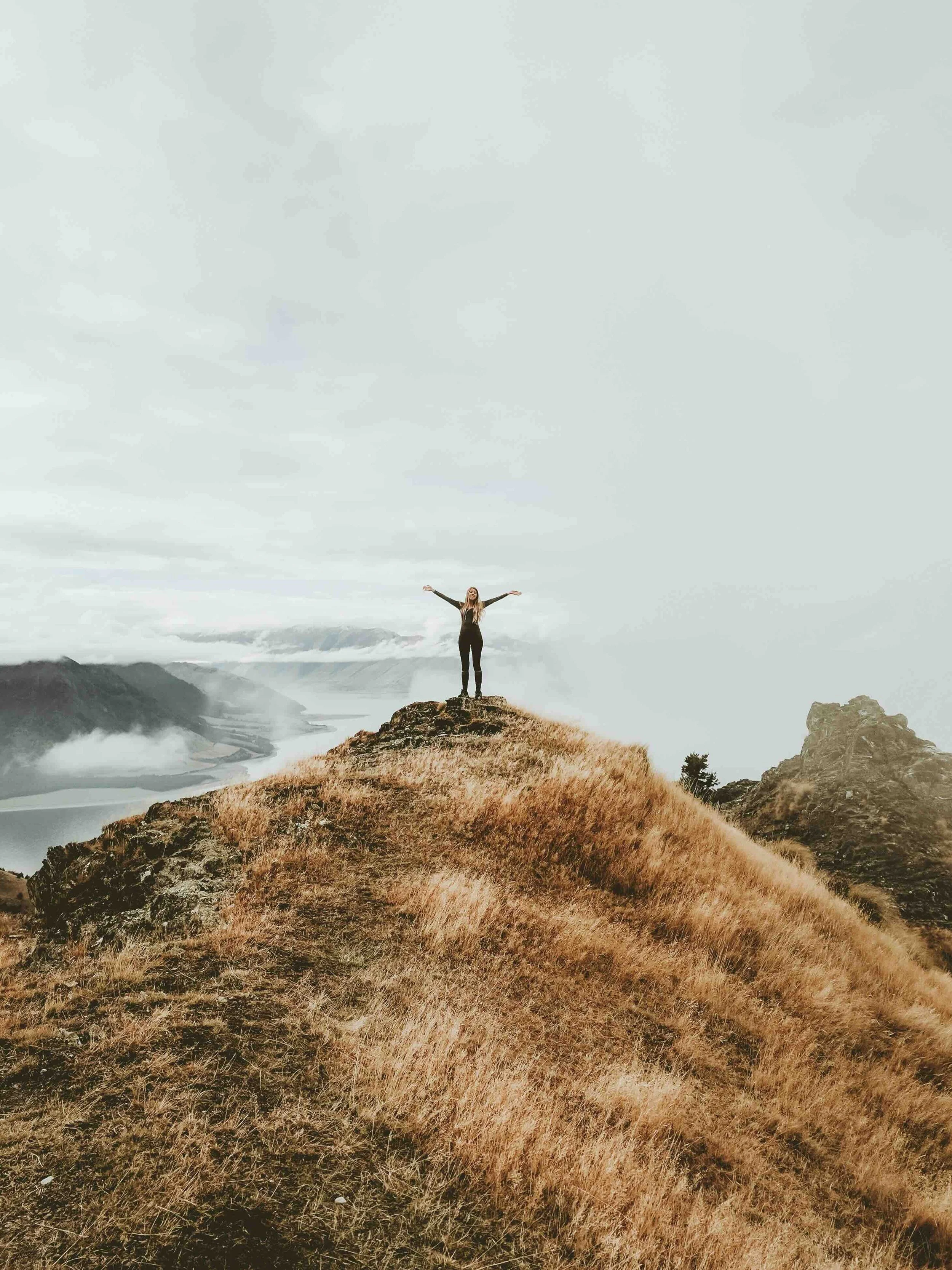 A person standing with arms outstretched on a grassy hilltop overlooking a body of water and mountains in the distance under a cloudy sky.